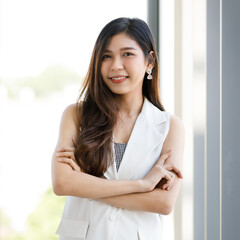 Portrait closeup shot of Asian young happy pretty confident brown hair female secretary wears white sleeveless long vest and blue jeans stand smiling look at camera in front glass window background