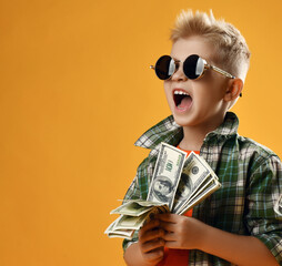 Cool blonde rich kid boy in brutal round sunglasses and checkered plaid shirt with the collar turned up, stands with fan of dollar cash in hands and screams isolated over yellow background