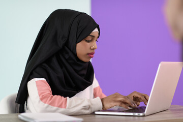 afro girl wearing a hijab thoughtfully sits in her home office and uses a laptop