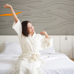 Asian woman in bathrobe rest on vacation, sitting in bed at home. Stretch after get up in the morning