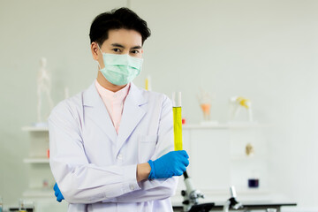 Scientific male researcher in white coat, green mask, and blue gloves standing with arms folded and holding a chemical tube of yellow liquid specimen in the chemistry analysis lab