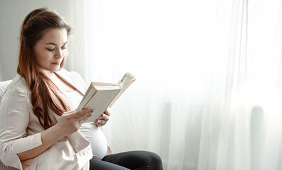 Young pregnant woman reading a book while sitting on the sofa at home.