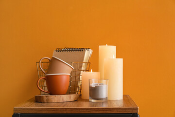 Burning candles, cups and notebooks on table against color background