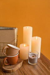 Burning candles, cups and notebooks on table against color background