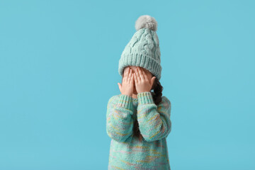 Little girl in winter clothes covering eyes with her hands on color background