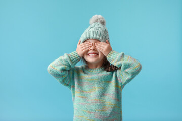 Little girl in winter clothes covering eyes with her hands on color background