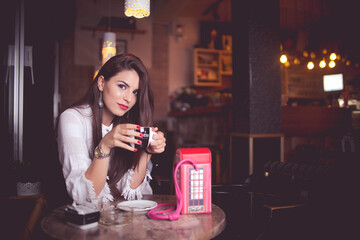 Beautiful brunette girl holding a coffee mug in a small dimly lit cafe in a fashionable trendy look