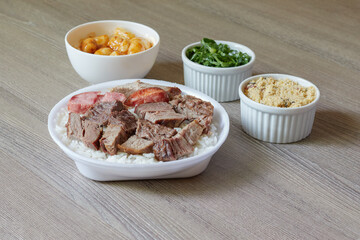 Marmitex with white rice, beans and barbecue with side dish of cabbage, crumbs and gnocchi on a textured wooden table.