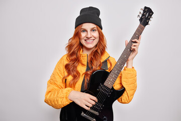 Fototapeta premium Beautiful redhead teenage girl learns to play electric guitar poses with musical instrument smiles pleasantly being pop musician wears trendry hat orange jacket isolated over white background