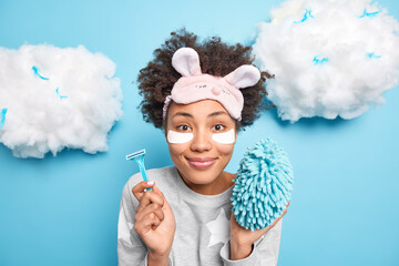 Pretty satisfied Afro American woman smiles gently undergoes skin care procedures wears slumber suit beauty patches under eyes to reduce wrinkles holds razor bath sponge isolated on blue wall