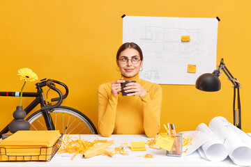 Thoughtful professional female architect drinks coffee poses at dekstop with building sketch in background has mess on office desk thinks about creating new project wears spectacles and turtleneck