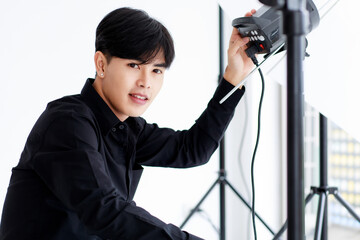 Portrait of an asian handsome male photographer or cameraman wearing casual black shirt, posing and looking into camera while setting up light equipment in a studio.