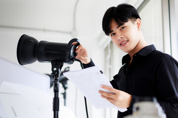 Portrait of an asian handsome male photographer or cameraman wearing casual black shirt, posing and looking into camera while setting up light equipment in a studio.