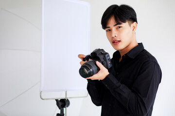 Asian handsome male photographer wearing casual black shirt, holding a camera, posing and sitting in a studio with light equipments.