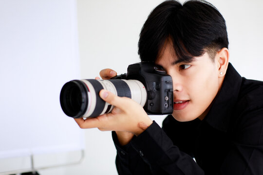 Asian handsome male model wearing casual black shirt with jeans, holding a camera, sitting on white background in a studio and posing while put his hand on hair.
