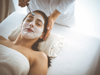 Beautiful brunette woman enjoying applying cosmetic mask with closed eyes comfortable and blissful. Relaxing treatment in medicine and spa center concepts