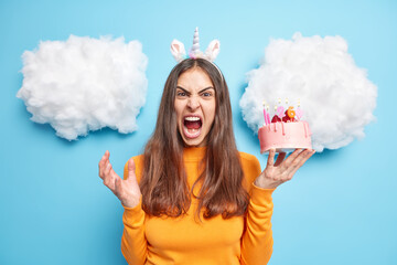 Outraged brunette woman screams angrily gestures actively screams loudly holds festive delicious cake wears orange jumper poses against blue background with white clouds overhead. Negative emotions