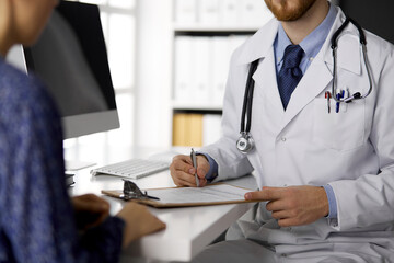 Unknown bearded doctor and patient woman discussing current health examination while sitting in sunny clinic, close-up. Medicine concept