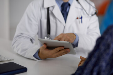 Unknown bearded doctor and patient woman discussing current health examination while sitting and using tablet computer, close-up. Medicine concept