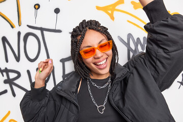 Energetic pleased dark skinned woman with braids expresses positive emotions dances carefree wears orange shades fashionable jacket poses against graffiti wall in public place. Teenagers lifestyle