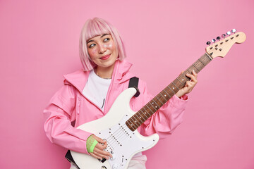 Fototapeta premium Pleased pensive female rock musician play white electric guitar perform popular song enjoys musical holiday wears pink jacket gloves stands indoor. famous artist has rehearsal before concert