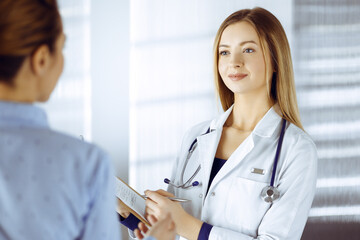 Young woman-doctor and her patient are discussing patient's current health examination, while standing together in a hospital office. Female physician is writing some marks, using a clipboard. Perfect