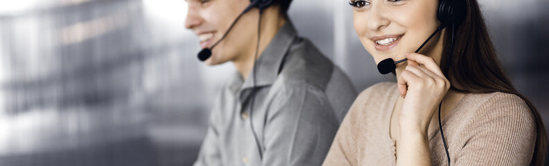 Two young people in headsets are talking to the clients, while sitting at the desk in an office. Focus on woman. Call center operators at work