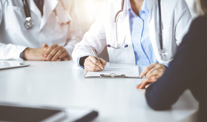 Unknown woman-doctor with male colleague are consulting patient woman while sitting at the desk in sunny clinic, close-up. Covid 2019