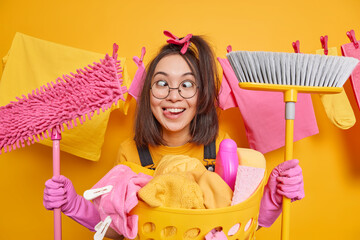 Asian woman makes funny grimace crosses fingers sticks out tongue holds broom and mop stands near laundry basket busy doing housework isolated over yellow background with clothesline behind. - obrazy, fototapety, plakaty
