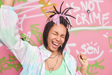 Overjoyed hipster girl dark skinned has combed braids foolishes around laughs and dances poses against colorful drawn graffit wall expresses positive emotions. Colored background. Street artist