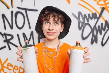 Handsome thoughtful curly haired teenager concentrated above holds two paint cans creats graffiti...