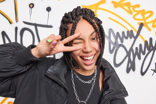Young positive graffiti artist makes peace gesture over eye smiles broadly dressed in black jacket and metal chians around neck poses against colorful drawn street wall. Modern generation concept
