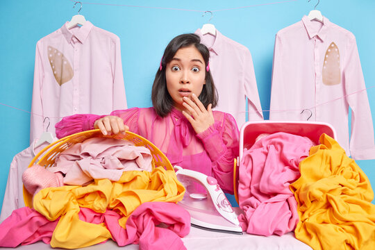 Image of shocked astonished female chambermaid looks stunned as has much work to do poses near laundry baskets uses electric iron works in hotel irons linen. Woman at dry cleaners. Ironing concept