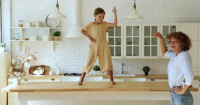 Vivacious little cute girl perform cool funky dances on dining table having fun together with young carefree mother in cozy modern kitchen on weekend. Home hobby, active quarantine with kids concept