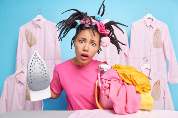 Surprised indignant dark skinned woman has funny hairstyle poses with stack of laundry going to iron does domestic work poses near ironing board against blue background washes linen uses detergent
