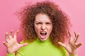 Portrait of angry woman frowns face exclaims annoyed makes car claws keeps mouth wide opened has curly bushy hair wears green turtleneck expresses negative emotionsn poses indoor. Anger concept