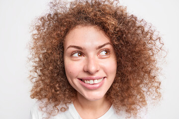 Headshot of good looking cheerful curly girl gazes away with daydreaming expression has healthy skin natural frizzy hair smiles broadly poses in studio against white background. Happy emotions