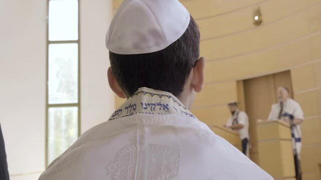 Bar Mitzvah boy with Talit and Kipa at synagogue. Jewish Kid with yarmulke having his ceremony in Israel led by a Rabbi, Hebrew Torah words on white  Talit, Tefillin. Judaism reform ceremony Holyland
