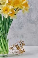 Bouquet of yellow daffodils flowers in glass and small dried decorative branchesstand on white wooden surface against gray concrete background. Spring concept, postcards, home decor, quotes.