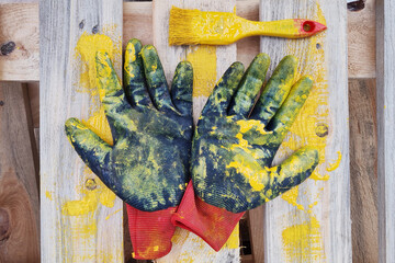 Black protective gloves and paint brush smeared with yellow painted lies on wooden pallet.