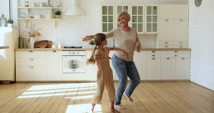 Cheerful active 60s granny little girl adorable grandkid listen music dance barefoot on warm wooden floor with underfloor heat system in cozy kitchen, full length. Funny activity, home hobby concept