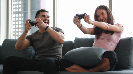 Caucasian couple enjoy playing on streaming game console on dark grey sofa in the living room. Console game fan activity at home concept.