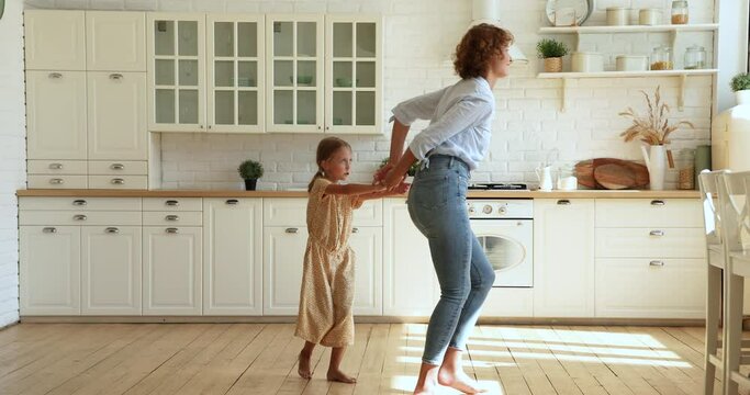Woman and small daughter listen music dancing indoor, active mom hold in hands lifting kid spinning enjoy funny playtime on weekend in cozy modern sunlit kitchen. Fun, happy homeowners, tenant concept