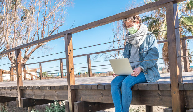 Spanish woman wearing a facemask with a laptop in her lap while sitting on a wooden bridge