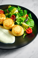 Salted cottage cheese pancakes with sour cream, baked cherry tomatoes and salad, served in a black plate on a marble background. Delicious and healthy breakfast. Ukrainian syrniki