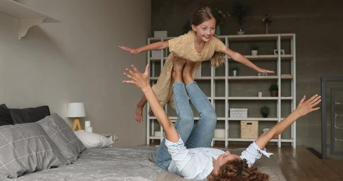 In cozy bedroom active strong mother lying on comfy bed holding lifting small daughter on legs, child spread arms like plane wings imagines like fly in air. Sportive family, yoga practice, fun concept