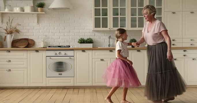 50s granny dance barefoot waltz with little graceful granddaughter in kitchen. Multi generational dancers in chiffon skirts hold hands enjoy active weekends together. Family bond, home hobby concept