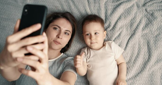 Mother taking selfie with baby boy using mobile phone. Woman and child lie on the bed and make video call using smartphone. Slow motion