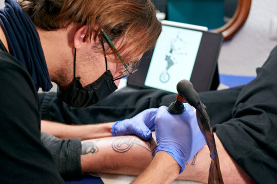 the tattoo artist in blue gloves looking at a tablet and doing a tattoo on the leg of a male in a studio