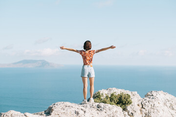 Rear view of brunette young woman standing with raised arms on the edge of a cliff  and enjoying of freedom on sunny day in summer vacations.